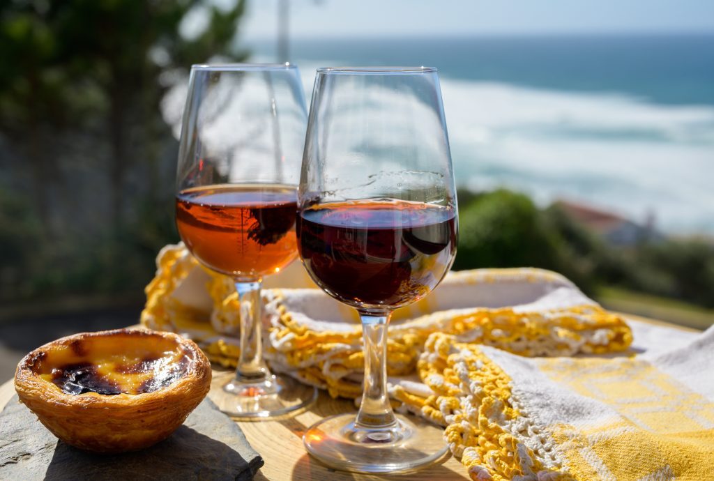 Portugal's traditional food and drink, glass of porto wine and muscatel de setubal, sweet dessert Pastel de nata egg custard tart pastry served with view on blue Atlantic ocean near Sintra in Lisbon area, Portugal