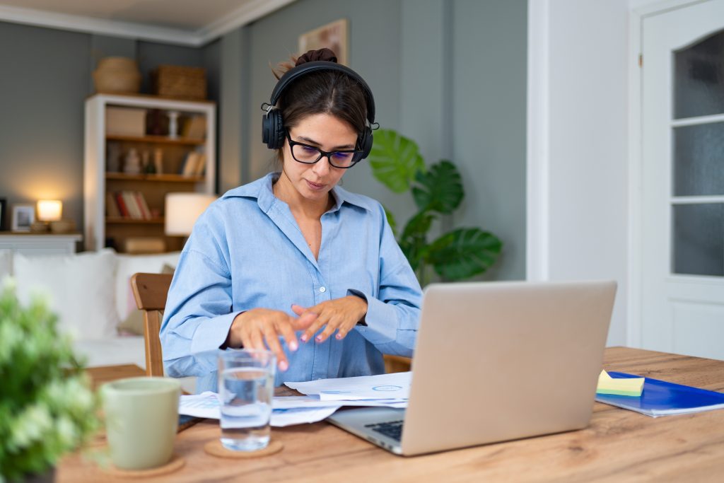 Frau im Home Office mit Laptop auf dem Tisch, Kopfhörern und blauer Bluse.