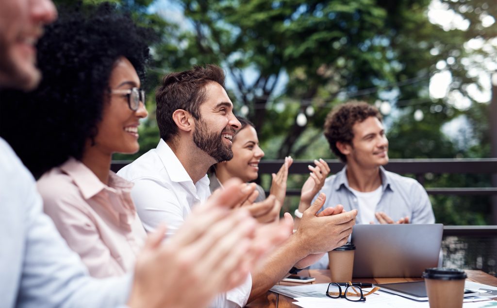 Team applaudiert in einem Meeting, draussen am Tisch mit Natur im Hintergrund.