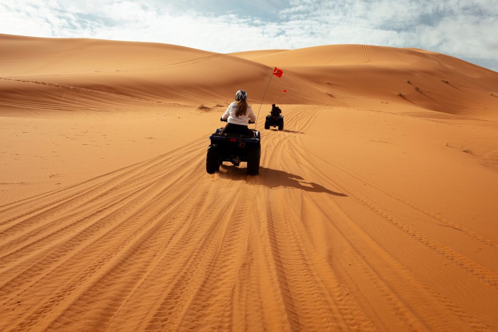 Quads fahren durch die Sahara-Wüste und wirbeln Sand vor den goldenen Dünen auf – ein abenteuerliches Erlebnis für Team Retreats, Incentive-Reisen und Wüstenaktivitäten in Marokko.